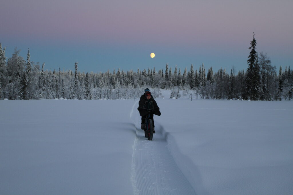 [Alt-teksti: Kaksi pyöräilijää ajaa keskelle lumimaisemaa auratulla pyöräväylällä kuun loistaessa talvisessa hämärässä.]