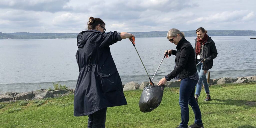 Three people by the lake collecting and picking up trash with litter picking sticks. 