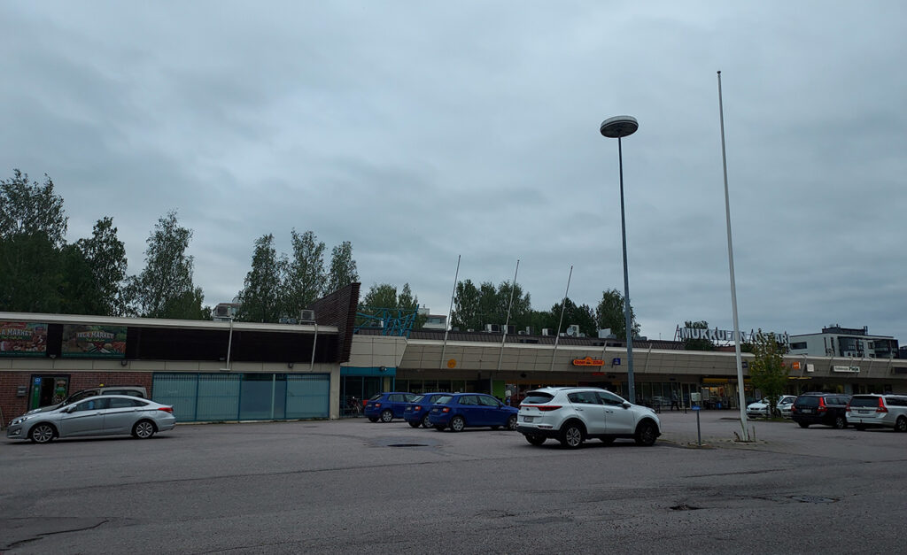 Mukkulan ostoskeskus pilvisenä ja harmaana päivänä. Autoja parkkipaikalla rakennuksen edessä.