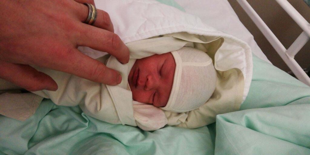 [Alt-teksti: Lämpimästi kapaloitu vastasyntynyt vauva nukkuu sairaalasängyssä ja aikuisen käsi kurottuu varovasti vauvan kasvoja kohti.]