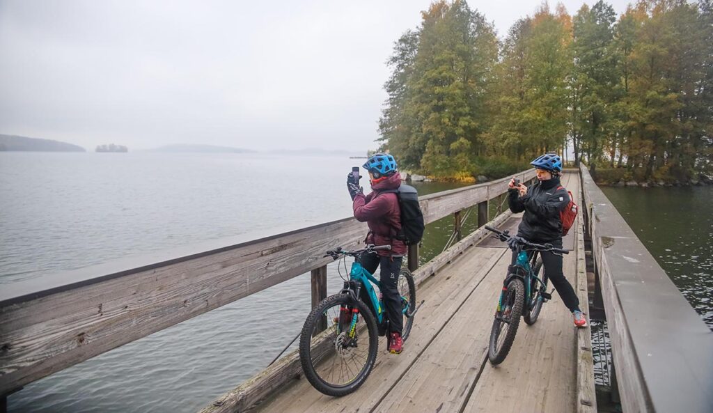 [Alt-teksti: Kaksi pyöräilijää puisella järven ylittävällä kävelysillalla ovat pysähtyneet ottamaan valokuvia hieman sumuisesta maisemasta.]
