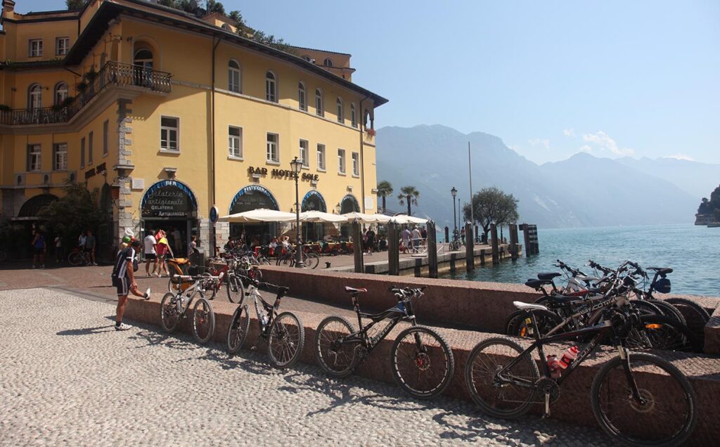 [Alt-teksti: Garda-järvi Italiassa. Vuorenrinteiden kehystämään järvimaisemaan on rantatielle rakennettu suuri kahvila, jonka vieressä on pysäköitynä toistakymmentä polkupyörää.]