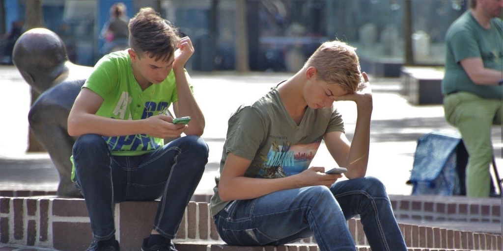 [Alt-teksti: Kaksi kesäisesti pukeutunutta nuorukaista istuu kaupunkipuiston reunamuurikiveyksellä älypuhelimiinsa syventyneinä.]