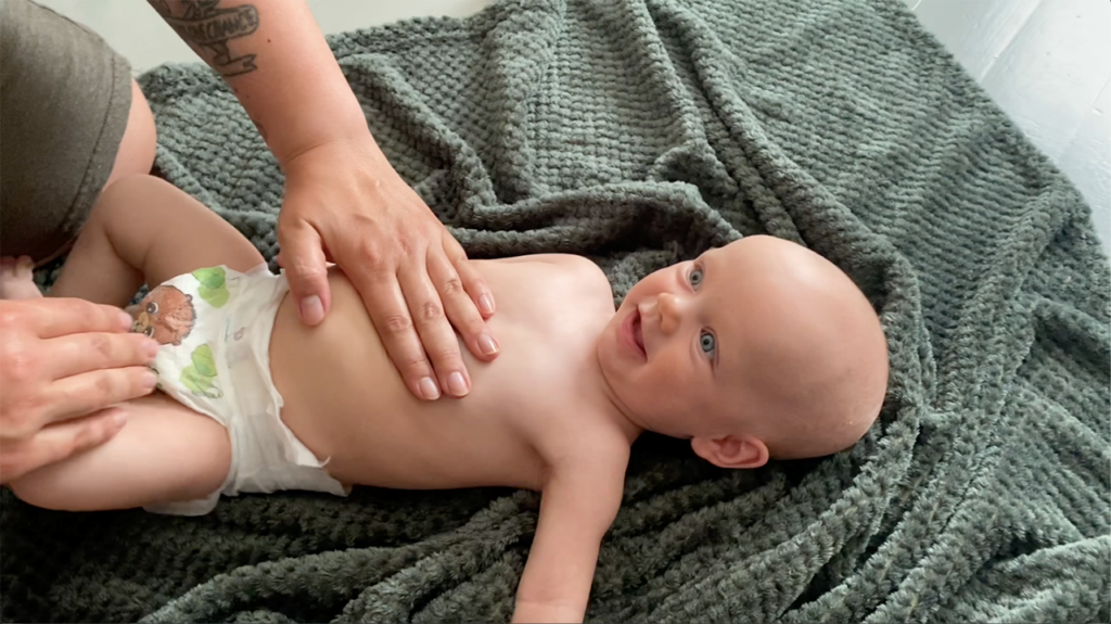 [Alt-teksti: Naurava vauva pelkkä vaippa yllään hoitopöydällä, aikuisen käsi vauvan vatsan päällä.]