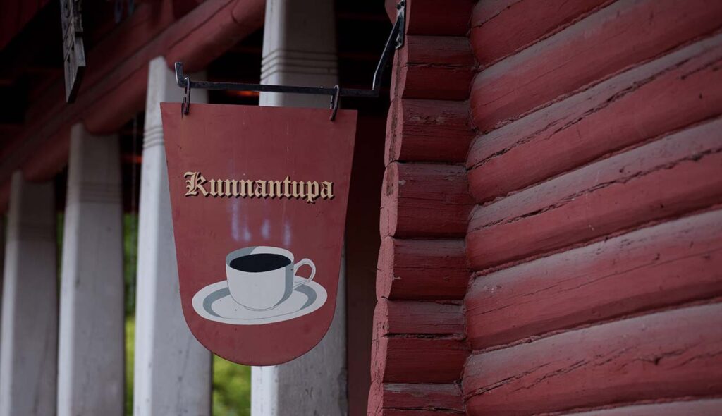 [Alt-teksti: Punamultamaalilla sivellyn hirsirakennuksen seinää, näkyvissä riippuva kyltti, jossa on kahvikupin kuva ja teksti Kunnantupa.]