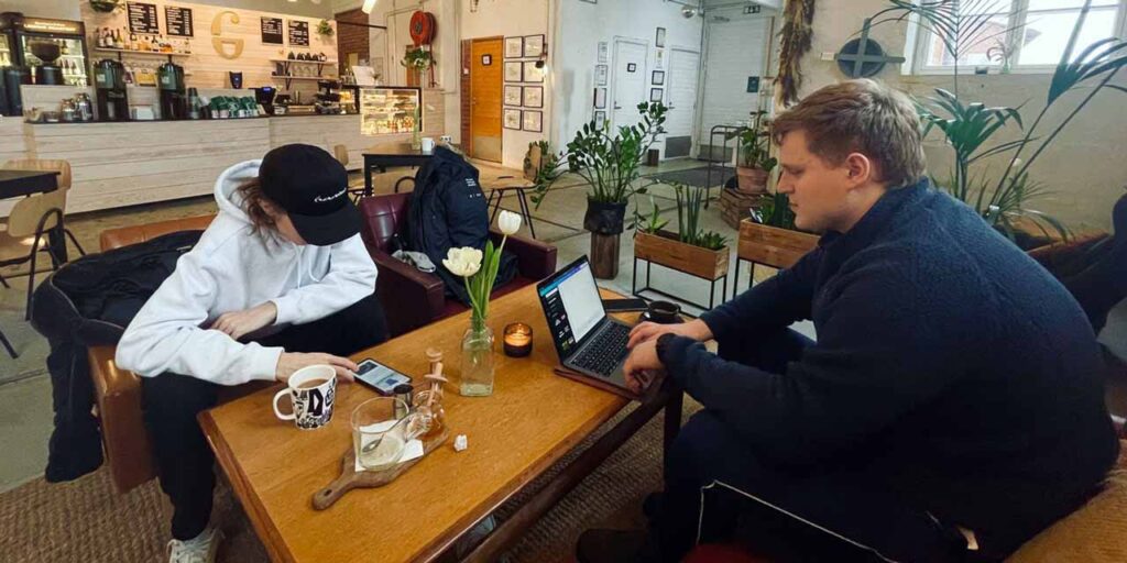 [Alt-teksti: Kaksi nuorta henkilöä istuu kahvilan pöydän ääressä, toinen näppäilee kannettavaa tietokonetta ja toinen tutkii älypuhelinta.]