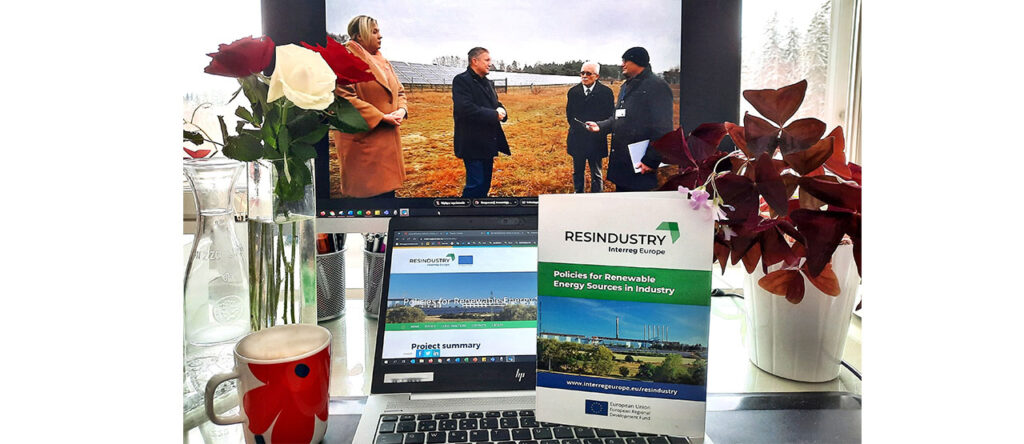 A photo of a computer and a big screen, on which the virtual site visit is just played. In front, there is the RESINDUSTRY project brochure standing. On both sides flowers are standing, on the left, there is a cup of coffee. 