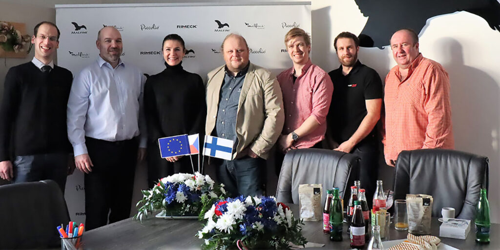 A photo of seven people standing next to each other, six men and one woman. In front of them, there is a table with flowers and Finnish, Czech and EU flags, as well as with some refreshments. In the background, on the right top, there is a logo of the company, a black eagle. 
