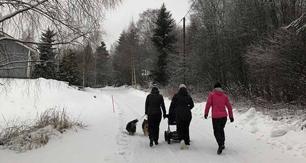 [Alt-teksti: Kolme nuorta naista kävelyllä lumisella maalaistiellä mukanaan lastenvaunut sekä talutushihnojen päässä kaksi koiraa.]