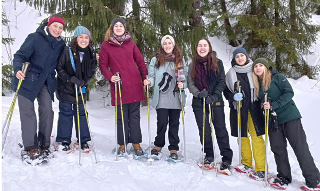 [Alt-teksti: Seitsemän naisen ryhmä lumisella rinteellä, takana suuria havupuita. Naiset seisovat rivissä ulkoiluvaatteissa, lumikengät jalassa ja sauvat käsissä, hymyillen leveästi.]