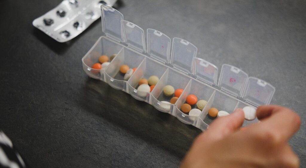 [Alt-teksti: Seitsenlokeroinen annostelija lokerot avoimina, lokeroissa on erivärisiä pillereitä, ja lääkeannostelua tekevän henkilön tablettia pitelevä käsi etualalla.]