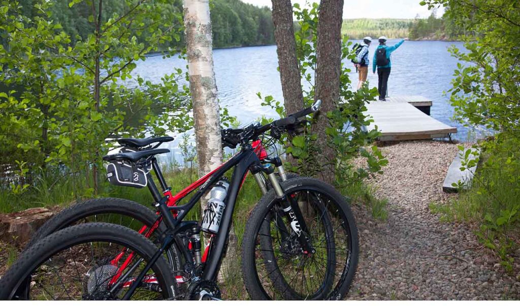 [Alt-teksti: Metsäinen lammenranta, jonka uimalaiturilla on kaksi retkeilijää. Heidän paksupyöräiset polkupyöränsä nojaavat puunrunkoon kuvan etualalla.]