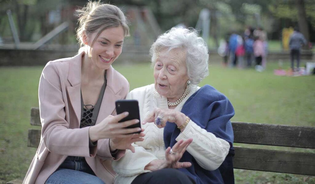 [Alt text: An elderly lady and a young woman sitting in the park, smiling and looking at mobile phone which is in youngster's hand.]
