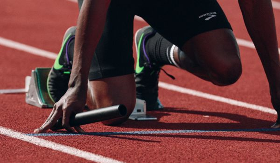 [Alt-teksti: Urheilija juoksuradalla lähtötelineissä. Urheilijasta näkyvät jalat juoksukengissä sekä käsi, joka pitelee viestikapulaa.]