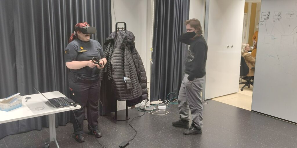 Two people standing in a room. The other one has VR headset on her head and controller on her hands. The other one stands and watches the ongoing VR simulation.