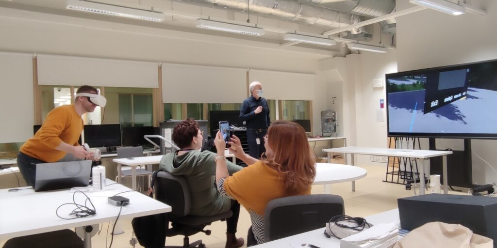 There are four people in a classroom. Two of them are standing and the other one has VR headset on his head and VR controllers on his hands. Two other people are sitting, the other one is watching a screen and the other one is taking a picture of the man who is wearing the VR headset on his head.