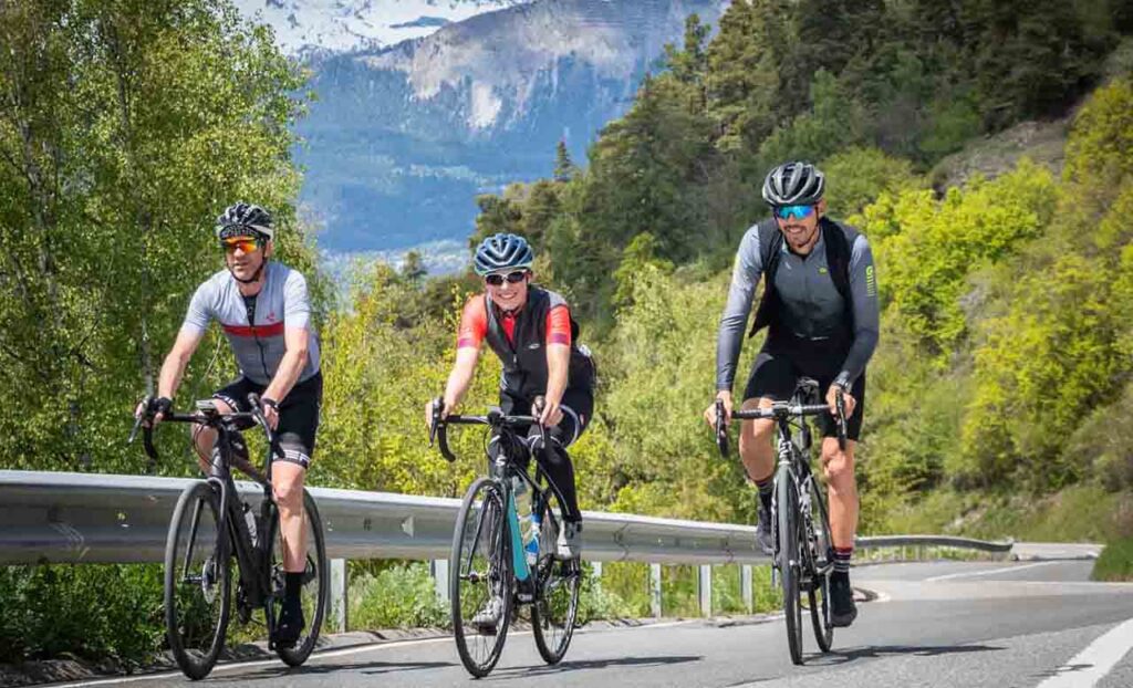[Alt-teksti: Kolme urheilullista pyöräilijää kesäisellä vuoristotiellä vehreiden maisemien ja vuorenrinteiden keskellä.]