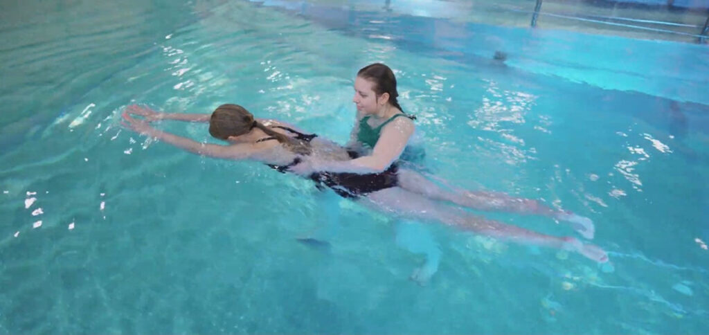 [Alt-teksti: Nuori uimari on altaassa kasvot vedessä ja raajat suorina, hänen vieressään avustava nainen tukee uimaria vyötäröltä.]