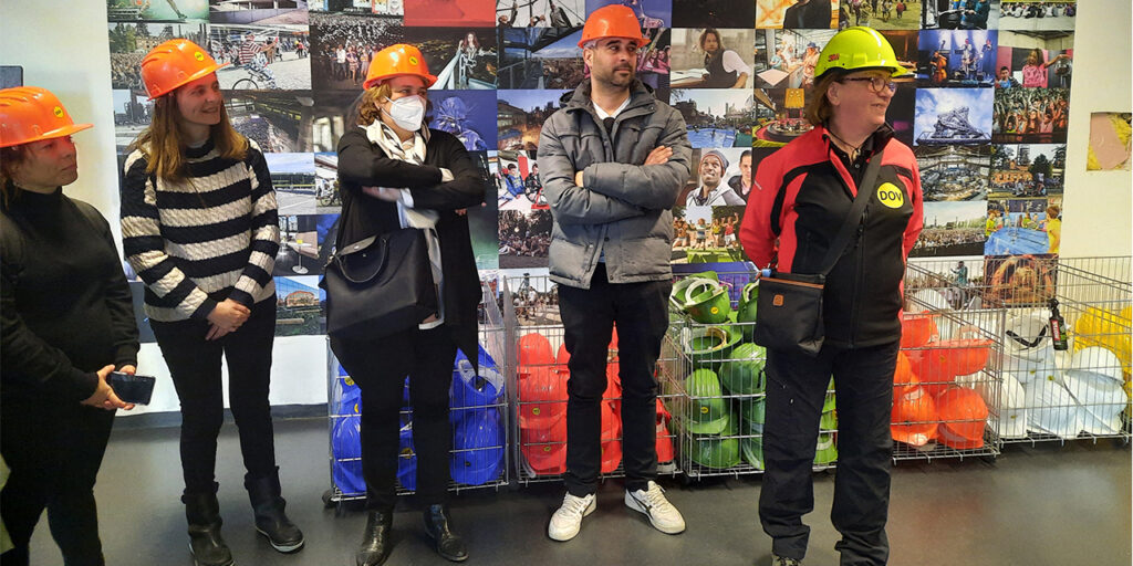 A photo of five people standing in a line and listening to one person, who is the guide. All people wear a safety helmet. At the background, there is a mosaic of colourful photographs and at the bottom, there are metallic baskets with spare helmets in different colours.