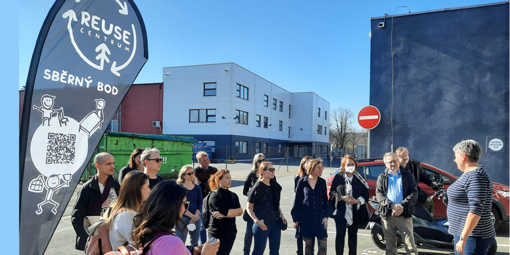 A photograph of a group of people listening and discussing with a local host. The picture is taken outside during a sunny day. On the left side, there is a sign of the visited place with a logo of the REUSE Centrum and other text and a QR code. At the background there is a high-volume green container and a white building.
