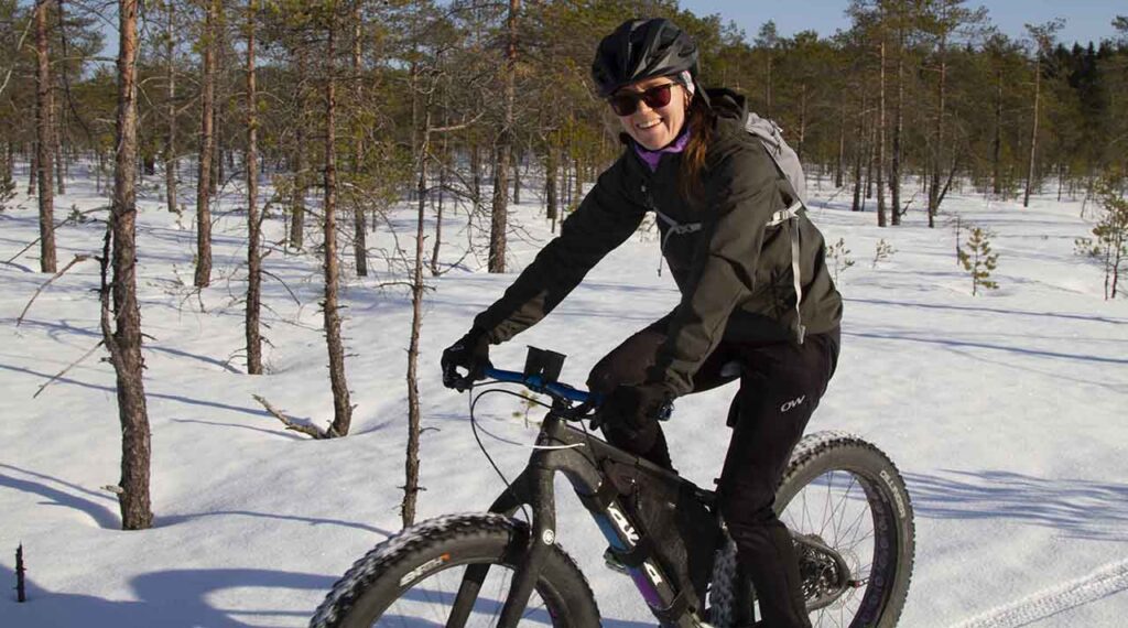 [Alt-teksti: Nainen ajaa paksurenkaisella maastopyörällä talvisessa metsämaisemassa hymyillen leveästi.]