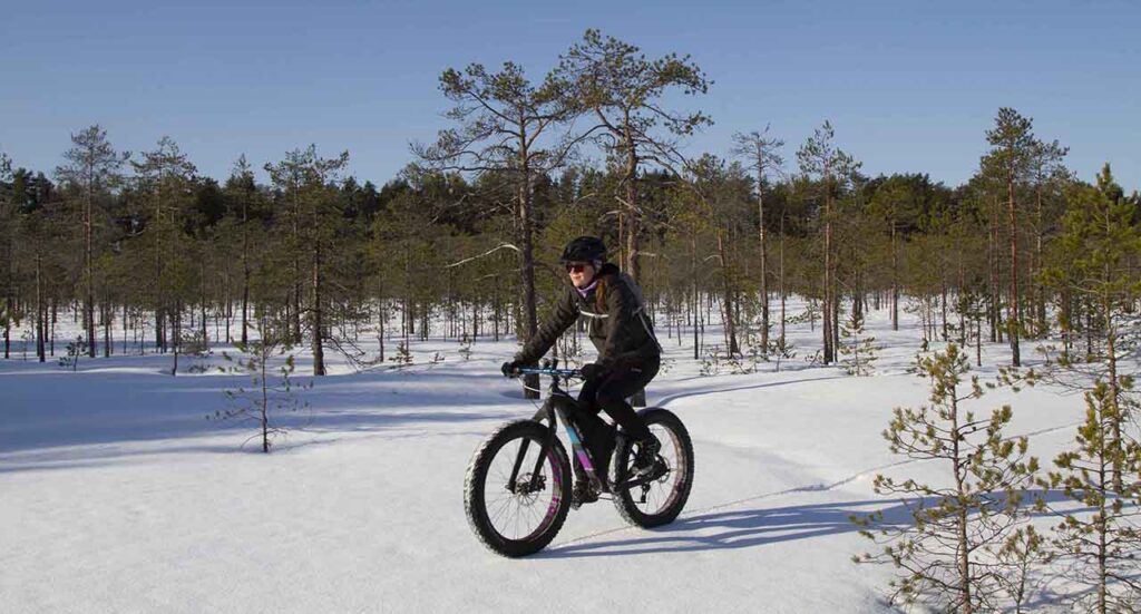 [Alt text: A sporty woman biking on snow near the forest.]