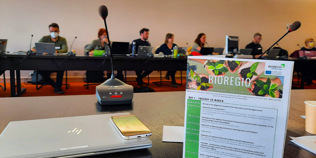 At the back of photo people are sitting in a meeting room and in front, there is a paper standing on the table saying BIOREGIO.