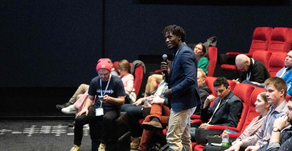 [Alt text: Young people listening in auditorium, one of them is speaking to microphone.]