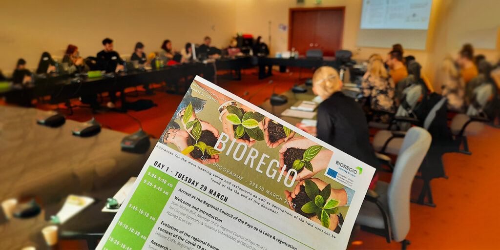 People are sitting around an oval table. There are microphones in from of every person. At the front, there is a paper with the agenda of Day 1 – Tuesday 29 March, with the title BIOREGIO and a project logo on the right corner of the paper agenda.