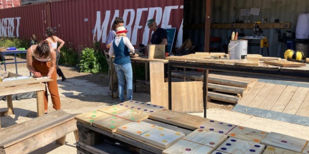 Five people working outdoors with fixing and painting wood objects. The weather is sunny.