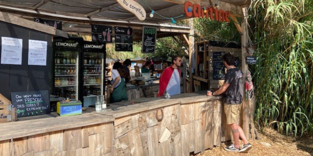 A bar desk outdoors with the text ‘bar’ and ‘cantine’ written with wood letters. Two persons standing by the desk.