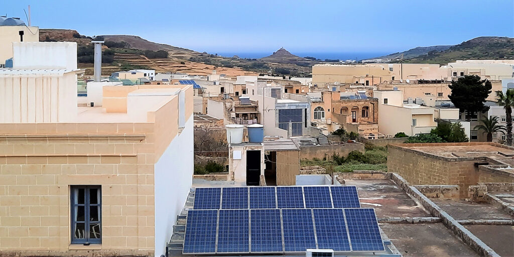 A view of sandy-coloured buildings, especially on their roofs, where solar panels are installed. In the background, there are mountains. 
