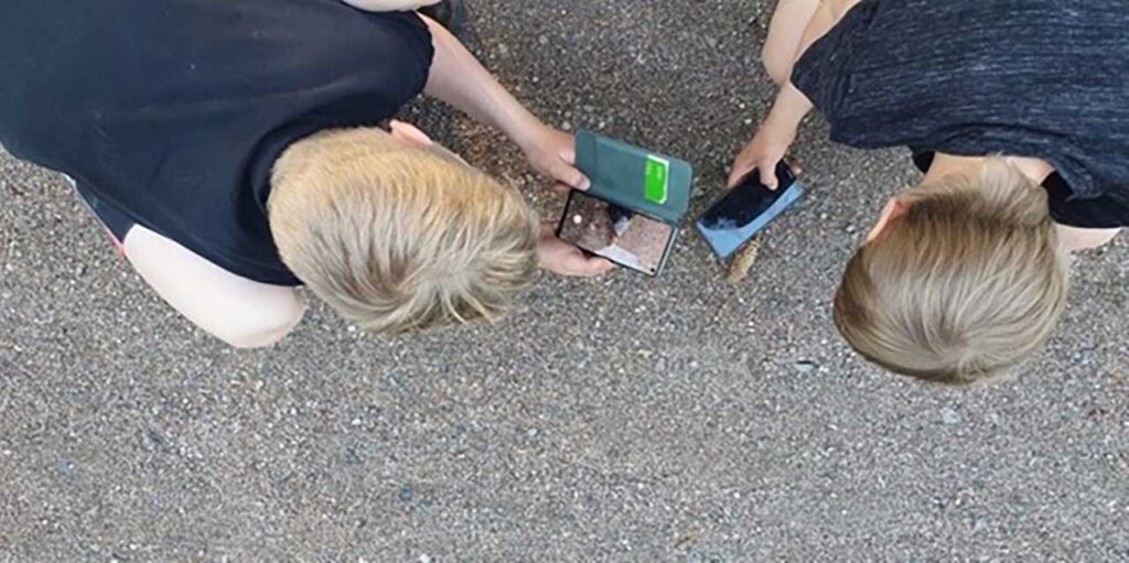 [Alt-teksti: Kaksi kouluikäistä lasta ulkona kyykkysillään pelaamassa mobiililaitteilla.]
