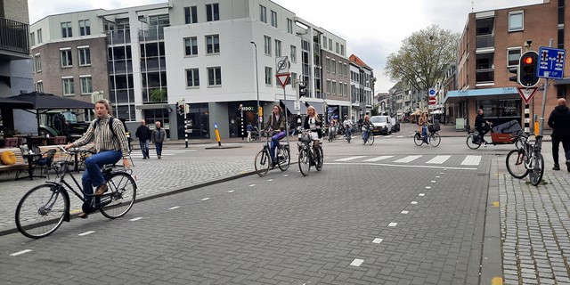 A picture of a road with separate bike lanes on both sides of the road in the city. There are also people cycling. In the background, there are buildings.