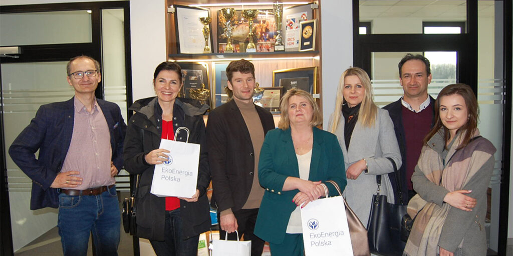 A photo of a group of seven people standing indoors in front of a glass vitrine with diplomas and awards. Three people carry a white paper gift bag with the EkoEnergia Polska and a logo. 