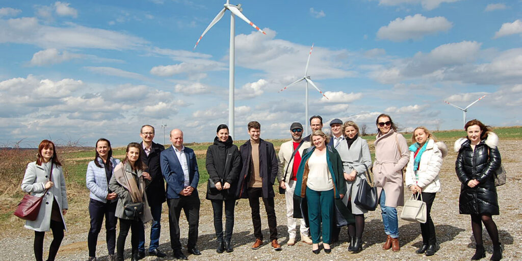 A photo of a group of people outside on a sunny day. In the background, there are three windmills. There are white clouds in the sky.   