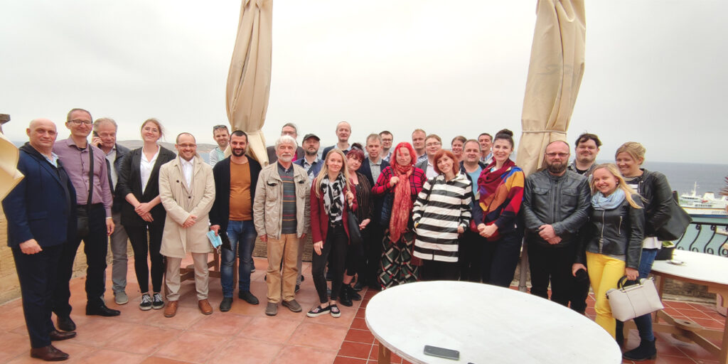 A group of people standing outside. On the front right side, there is a white round table. In the background, there are two unopened sunshades. 