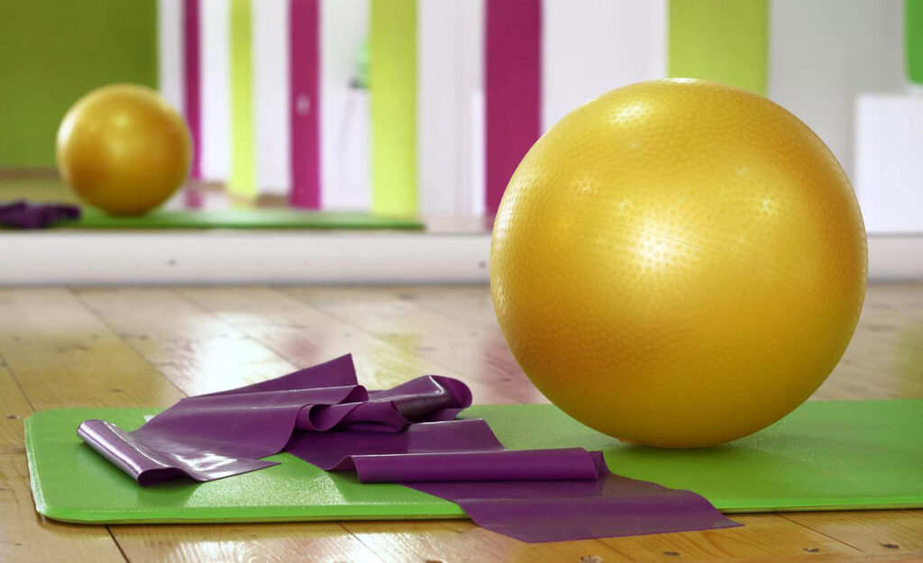 [Alt-teksti: Liikuntasalin puinen lattia, takaseinällä peili, lattialla on liikuntamatto, suuri keltainen jumppapallo ja violetti jumppakuminauha.]
