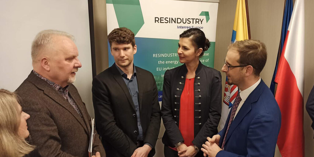 A picture of five standing people in formal clothes discussing something. Behind them, there is a roll-up with a logo of RESINDUSTRY, on the right back, there are flags.