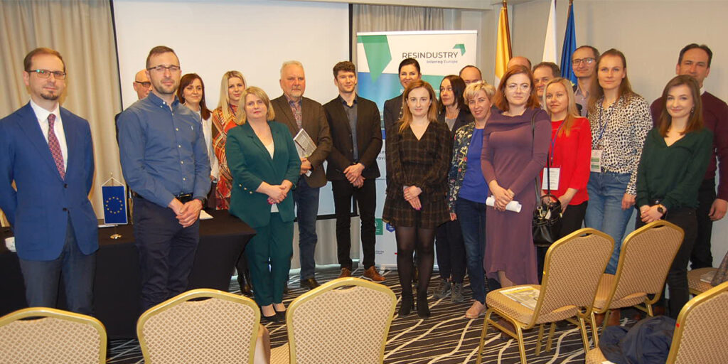 A photo of a group of people standing in a conference or meeting room. In front of the group, there are chairs organized in rows. At the back, a screen, roll-up with a RESINDUSTRY logo and some flags are partly visible.