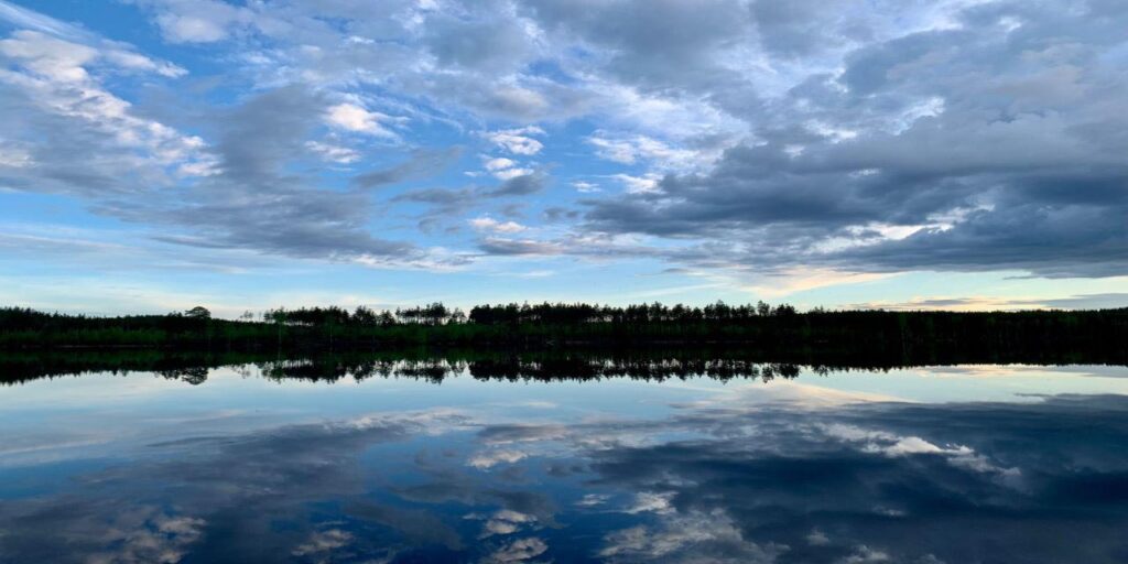 [Alt-teksti: Laaja järvinäkymä, peilityyni vedenpinta heijastaa taivaan pilviä, kaukana horisontissa metsänreuna.]