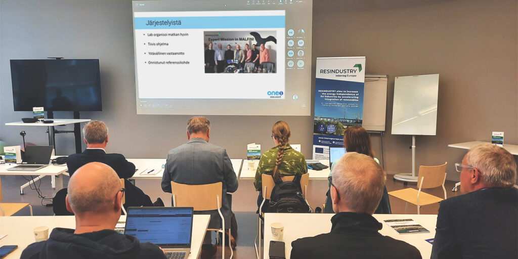 a group of people sitting indoors in a meeting room in two rows. They are listening to a presentation and looking at the big screen. On the screen, there is some text and a photo of seen people. On the right of the screen, there is a project roll-up standing with the title RESINDUSTRY.