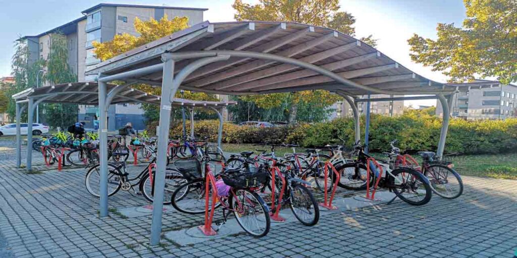 [Alt-text: Polkupyöriä pysäköityinä kahteen katokselliseen pyöräparkkiin, joissa molemmissa on punaiset runkolukitusputket. Taustalla on kerrostaloja ja puilta sekä pensaita.]