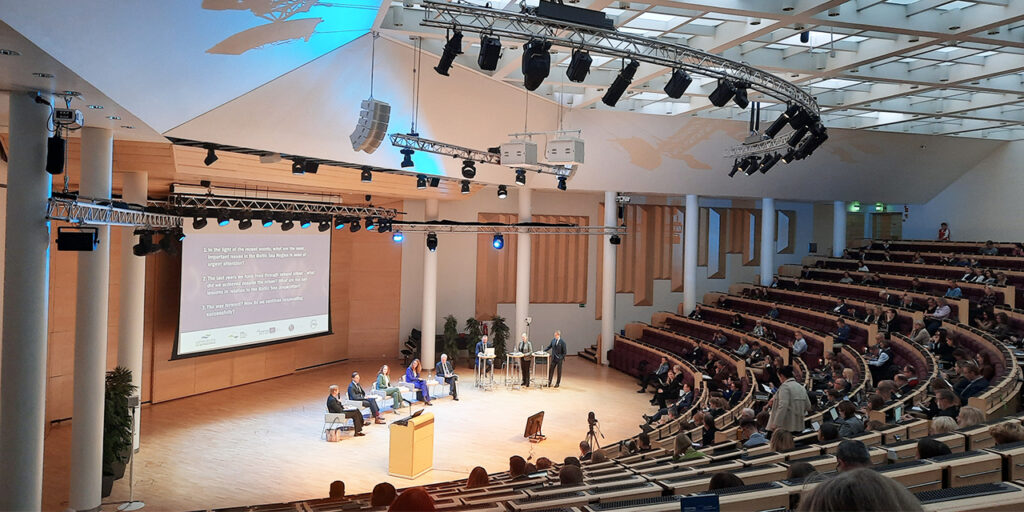 A view of a conference hall with people sitting in elevating seat rows. In the centre, there is a stage with five people sitting in chairs and three standing speakers. At the back of the stage, there is a big screen with some text. 