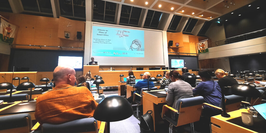 A photo of a meeting room indoors with rows of wooden tables and chairs and black table lamps. People sit on the chairs and listen to the speaker, who stands in front of a big screen. On the screen, there is a picture of a child holding a wooden plane and the text Citizens as Pilots of Smart Cities.