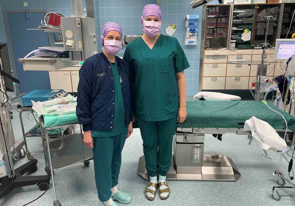 [Alt-teksti: pienehkö leikkaussali täynnä hoitovälineitä. Leikkauspöydän edessä seisoo kaksi sairaanhoitajanaista kasvoillaan suojamaskit ja päässä suojapäähineet.]