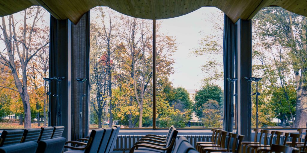 Kuusi riviä Alvar Aallon suunnittelemia 68-mallin tuoleja sekä nojatuoleja sivultapäin kuvattuna. Tuolien taustalla näkyy koko seinän levyinen ikkuna, josta näkyy ruskan värjäämiä puita. Tilan katto on vaaleaa puuta ja siinä on aaltoileva muoto.
