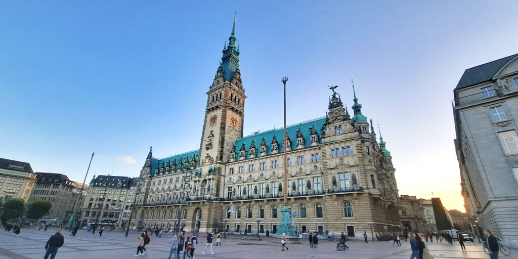 An outdoor photo of an old building serving as a city hall. It has a tower in the middle and a green roof. The building is located on a square, where people are walking in many directions.