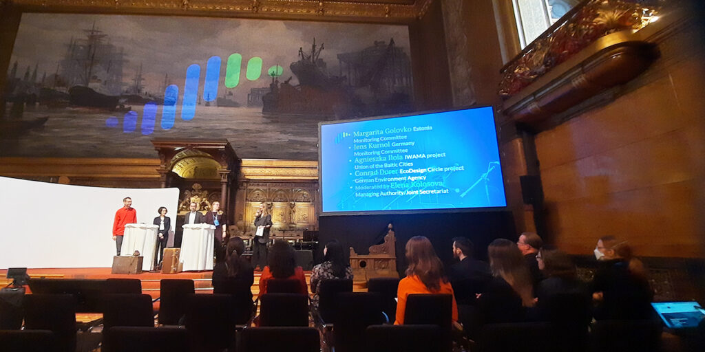 A photo taken indoors with a decorative interior and a big painting in the background. There is a colourful logo consisting of vertical lines projected on the painting. Under the painting, there is a stage, on which five people are standing. On the right side, there is a big screen with a list of speakers’ names. In the front, the audience is sitting on dark chairs facing the stage. 