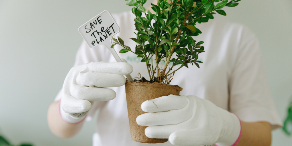 A person in a white t-shirt and gloves holding a pot with a green plant and a sign stating save the planet. 